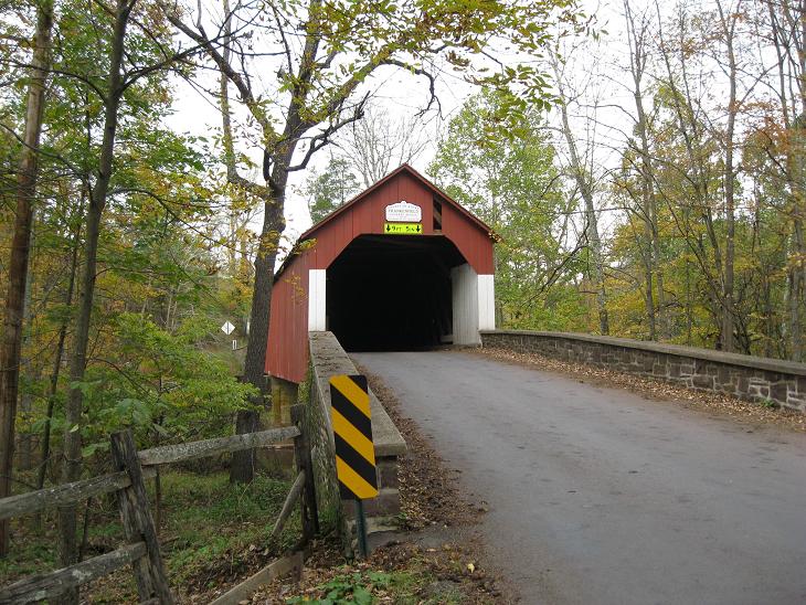 Red painted covered bridge (Frankenfield Covered Bridge) arching over Tinicum Creek, flanked by green trees and rural landscape.