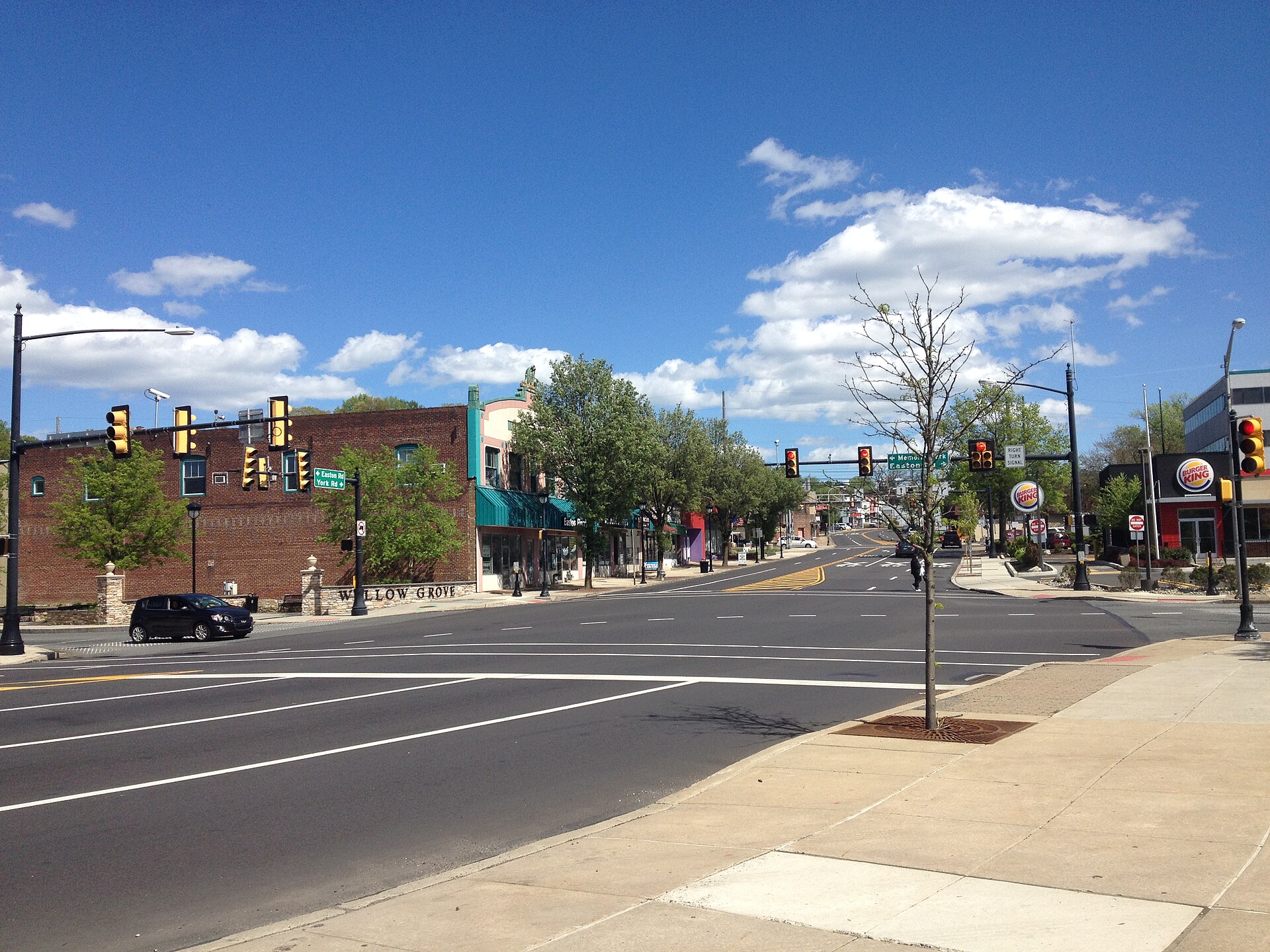 A wide view of a multi-lane roadway in Willow Grove, Pennsylvania, showing PA Route 611 (Easton Road) with traffic moving through a suburban corridor. A green highway sign and nearby intersection indicate Memorial Park and Easton Road, with roadside trees, utility poles, and commercial buildings lining the street under a clear sky.